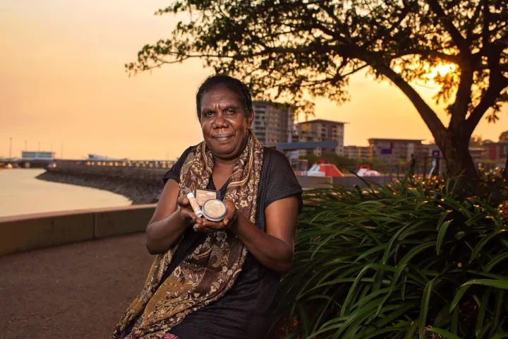 An Aboriginal woman in dark dress and patterned scarf sits next to a bush and holds three small containers in her hands. In the background is water, a wharf, a tree and buildings.