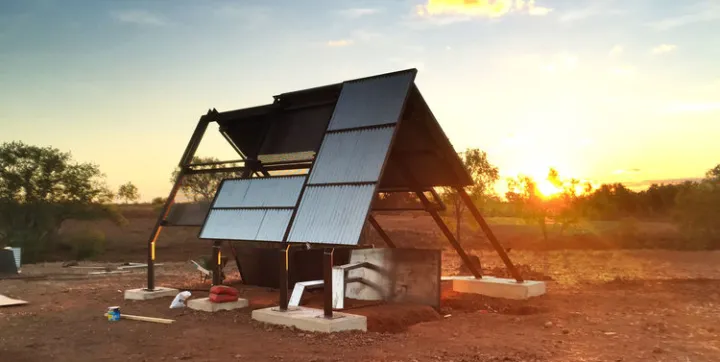 A structure of iron and metal sheeting stands on blocks of concrete. In the foreground is red soil and in the background are trees and a setting sun.