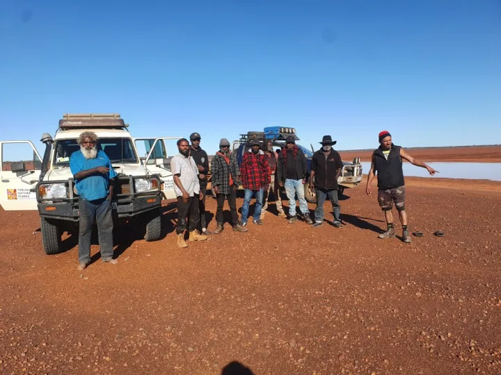 A group of men dressed in casual outdoor wear stand shoulder-to-shoulder on stony and ochre coloured soil. Behind them are two vehicles and in the background is a claypan and blue sky.