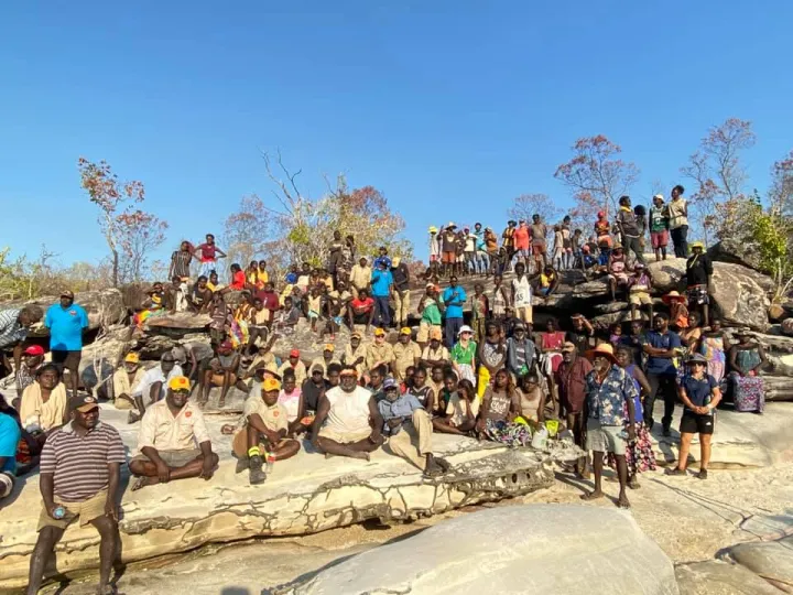 A large group of men, women and children wearing casual or work wear sit or stand on a rocky outcrop facing the camera. In the background are a few trees and a blue sky.