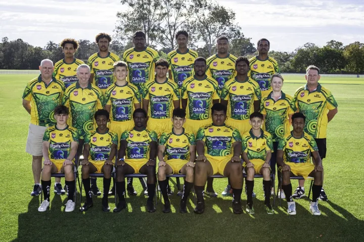 A group of mainly young men in sporting attire sit and stand on a grassed playing field in three rows. They each wear a blue, yellow and green jersey. In the background is green grass and trees.