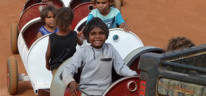 Young Aboriginal children sit in and stand next to a series of barrels mounted on wheels.