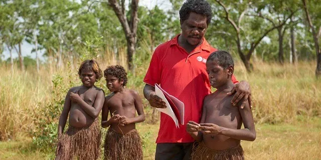 Two Aboriginal boys in grass skirts stand at left and behind an Aboriginal man in a red shirt and boy in a grass skirt in the foreground. The man holds some papers on which he and the third boy are focussed. In the background is long grass and trees.