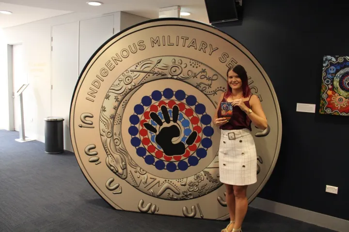 A woman in pale skirt and dark top stands next to a large replica of a coin, which features a hand in the middle of concentric circles, made up of black, red and blue dots. The coin is gold in colour. In the background are walls and a carpeted floor.