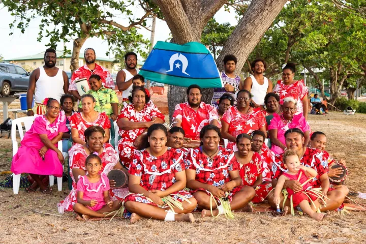 Group of people ranging from elderly to young children sit on the ground or seats or stand in front of a tree. On the tree is a blue, white and green flag. In the background are more trees, a car, a house and more people.