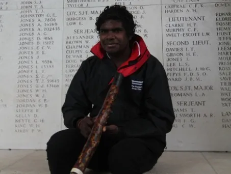 Elijah Douglas, holding his didgeridoo, sits in front of a wall of remembrance.