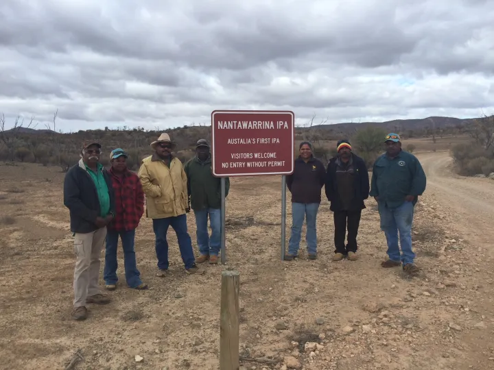 Seven Aboriginal men and women dressed in warm clothing stand next to a red sign just off a dirt road. In the background are bushes, trees and hills. The sign says Nantawarrina IPA – Australia’s first IPA – Visitors welcome – No entry without permit.