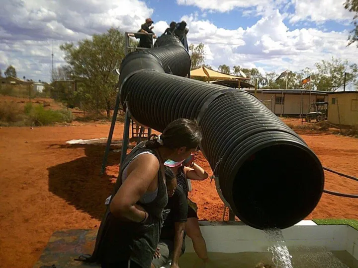 A long and very wide black pipe snakes from a stand downward to a pool. On the stand are several people and at the pool are a few more. In the background is red soil, trees and buildings.