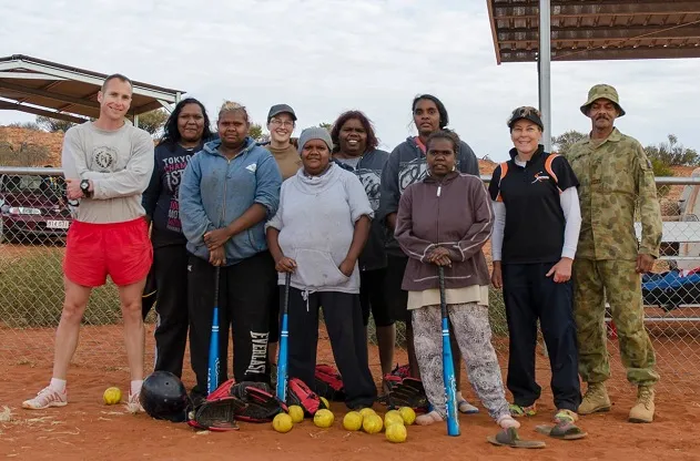 Indigenous women with non-Indigenous army men and women wearing various clothing types and some holding softball bats. There are softballs on the red earth in front of them.