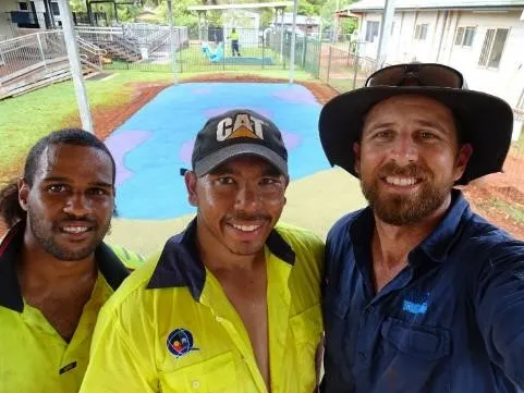 Three men, two in yellow work-wear shirts and one in a blue shirt stand close to camera. In the background is a flat area covered with blue and pink rubber. Surrounding this is bare soil, grass and fences. There are buildings at right and left and behind.