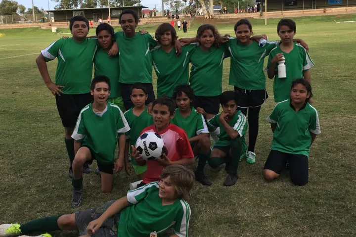 On a grass field is a group of 14 Aboriginal youth in green soccer jerseys and black shorts. The back row is standing, the middle row is kneeling and one young man lays down in front of the rest. One young man is in a red shirt and holds a soccer ball. In