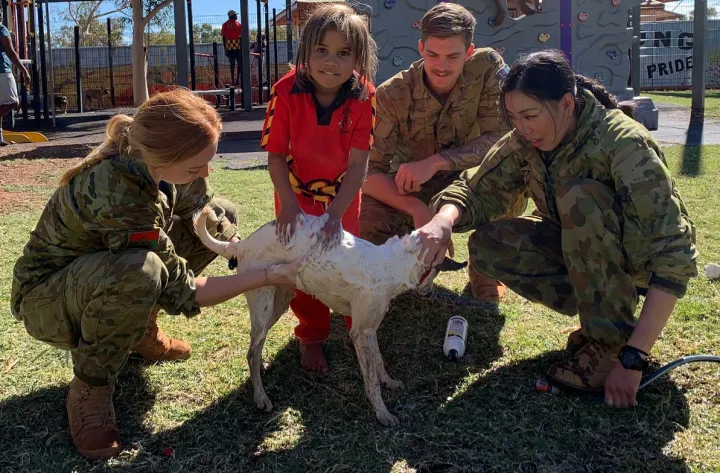 Young Aboriginal girl in red school uniform and three soldiers (2 women and 1 man) surround a white dog on a grassed area. One soldier holds a hose. In the background is a playground.