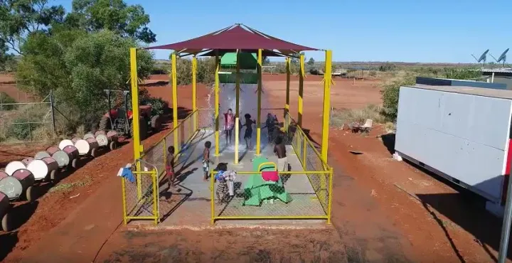 Several Aboriginal children play on a concrete slab under sun shade amongst various water features. On the left is a small train with carriages made from barrels and on the right is a building. In the distance is open ground, more structures and bushland