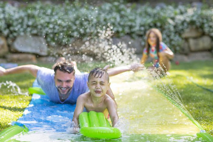A small child holding a green inflatable doing down a slip ‘n’ slide in the back yard. Behind his is an older man on the slip and slide. In the background is a young girl holding the hose on the waterslide