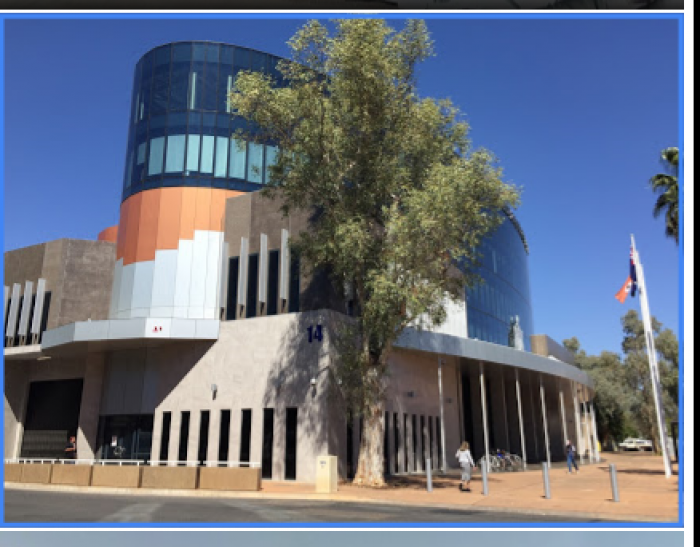 Blue, orange, white and beige building with tree in front and footpath with pavers in front of that. Aboriginal flag at right.