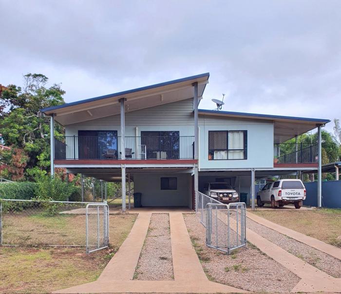 In the foreground are two driveways divided by a wire fence leading to a house divided into two parts and each with two levels. On the right are two vehicles. In the background are trees and a cloudy sky.