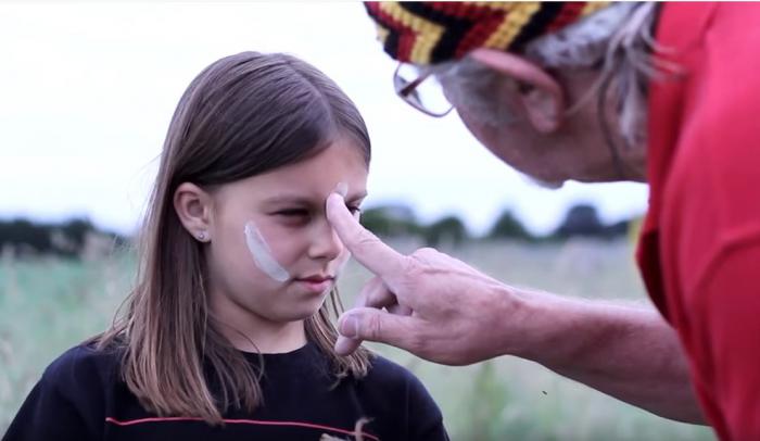 The face of a young girl is painted with white stripes by an elderly man in red shirt and head wear in black, red and yellow.