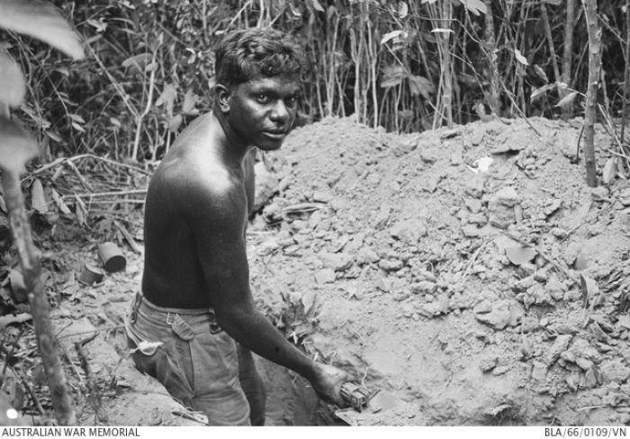 A young man wearing shorts but bare chested holds a shovel in his hand while he stands in a trench. In front of him is a pile of soil and beyond that is thick foliage.