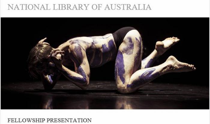 Main painted with purple Indigenous designs on hands and knees on a smooth black floor. Above and below him are the words National Library of Australia and Fellowship Presentation.