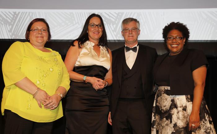 Four adults stand in a row: woman in yellow top, woman in white and black dress, man in tuxedo and woman in black top and black and white mottled skirt. All wear glasses and stand in front of a two toned wall.