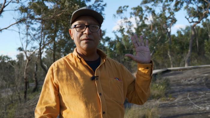 Elderly man in yellow shirt and peaked cap stands with left hand raised. In the background are trees and bare ground.