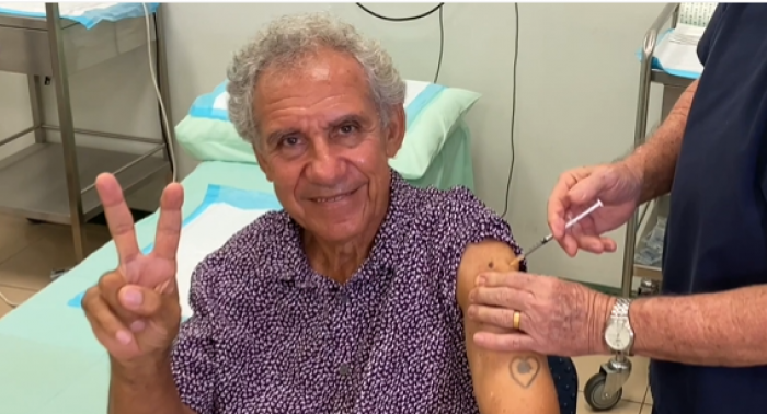 A man in a purple and white spotted shirt is sitting next to a medical bed with two fingers in a peace sign. A second man in blue scrubs is putting a needle in the first man’s arm.