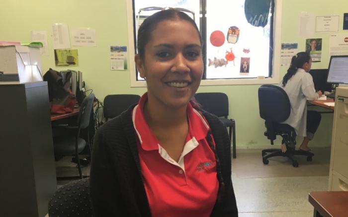 Young Aboriginal woman in red shirt sits in an office with equipment and her colleague in the background. The floor is grey and the walls green in colour.