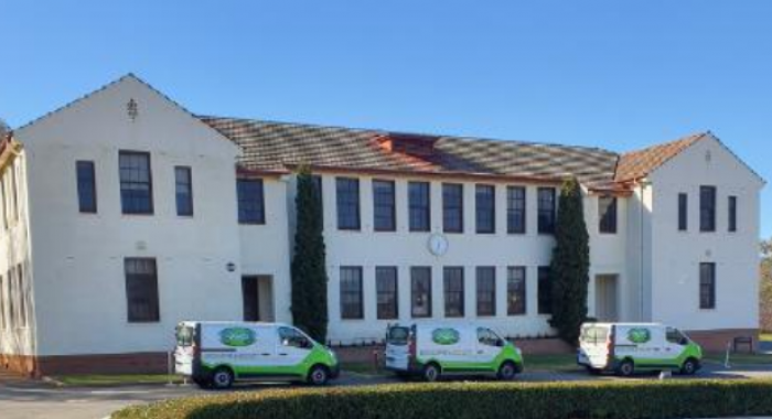 Three white and green vans line up on the roadway in front of a large white building with lots of windows and a red tile roof.