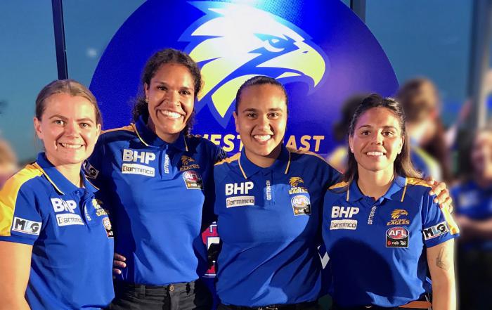 Four Indigenous women in blues shirts stand shoulder to shoulder. They wear blue and yellow shirts. Behind them is a sign with a logo featuring an eagle’s head.