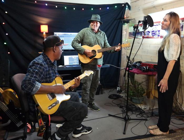 Three people in a small space, wearing casual clothing, are making music. Two men play guitars while a woman stands at a microphone. In the background is recording equipment.