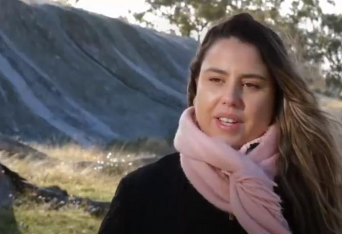 A head and shoulders view of a woman with long hair and wearing a black top and pink scarf. In the background is grass, a large long rock and trees.