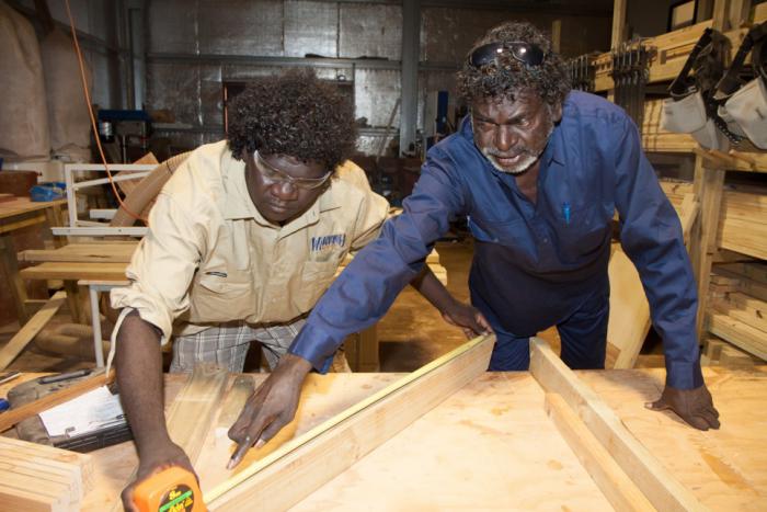 Two Indigenous men working at a wood working bench with the elder man directing the younger