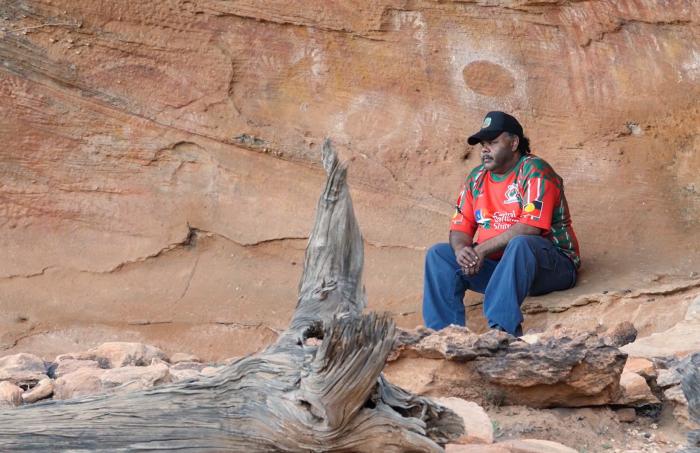 Aboriginal man in red shirt, blue jeans and a black cap sits on a rock ledge behind which are cave paintings. In front of him is a fallen log.