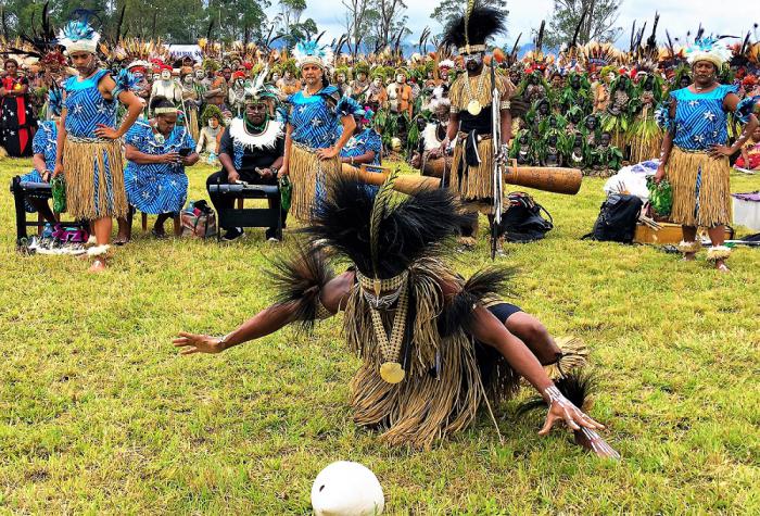 A dancer in traditional Torres Strait Islander dance wear squats on grass looking at a white round shaped object. In the background are many people dressed in dance costumes, trees and sky.