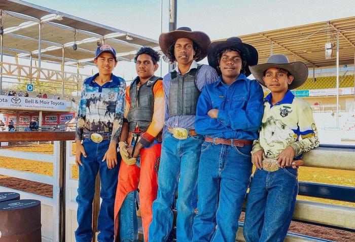 Five boys stand side by side leaning against a wooden fence. Behind them is a rodeo ring. Each of the boys is wearing jeans and a belt or work pants and collared work shirts. The boy on the left is wearing a cap and four of the boys aren't.