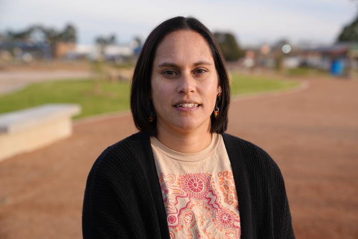 A woman with dark shoulder length hair and wearing a black top over a colourful top with traditional designs faces camera. In the background is brown soil, green grass and some buildings.