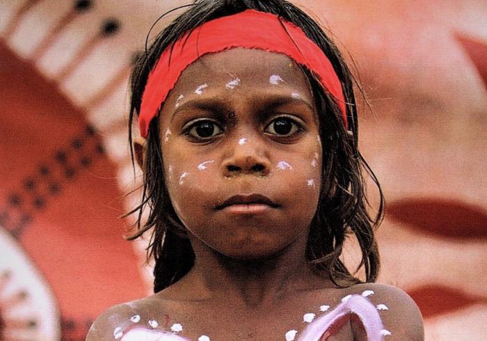 Aboriginal boy in body and face paint standing in front of Aboriginal artwork