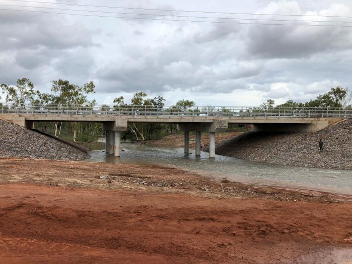 In the foreground is ochre coloured soil behind which is a concrete bridge spanning a flowing creek and in the background are trees and a cloudy sky.