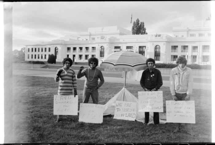 Four men in casual wear stand on grass and either side of a beach umbrella. In front of them are small hand written signs. In the background is a road and then a white building with many windows.