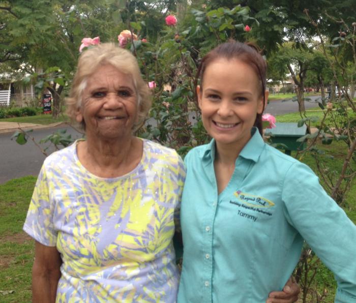 Elderly Indigenous woman in yellow and grey dress stands with arm around young Indigenous woman dressed in aqua coloured shirt in front of a flowering bush in a suburban street.