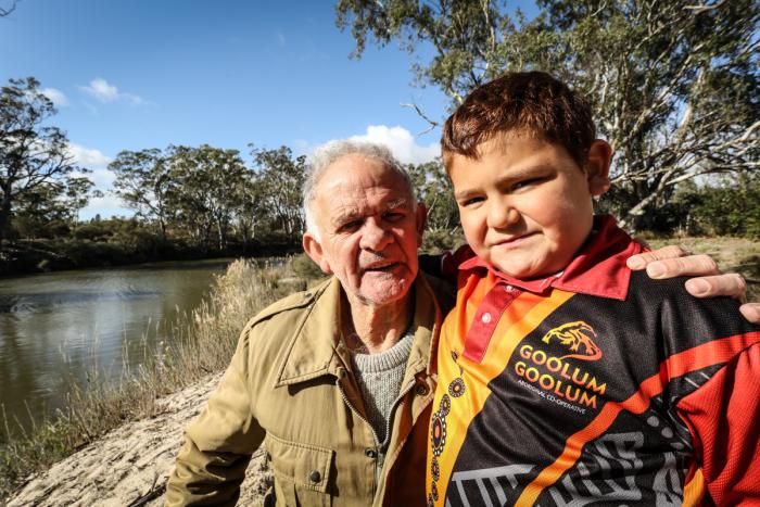 An Aboriginal man in brown jacket and boy in red, black and yellow shirt, stand near a river behind them. Also in the background are trees, grass and a blue sky.
