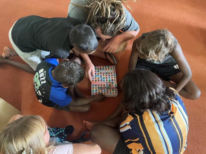 The arrival of the Warlpiri Encyclopaedic Dictionary in Yuendumu, Northern Territory. Five children and an adult are bent over the Warlpiri Encyclopaedic Dictionary. The cover of the dictionary has photos of people who have contributed to it.