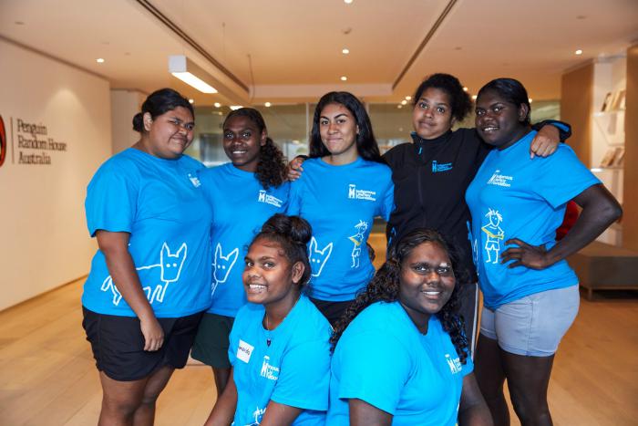 Seven young Aboriginal women, most wearing blue t-shirts stand in a group in a room with a board floor and a book case in the background. At left on the wall are the words Penguin Random House Australia.