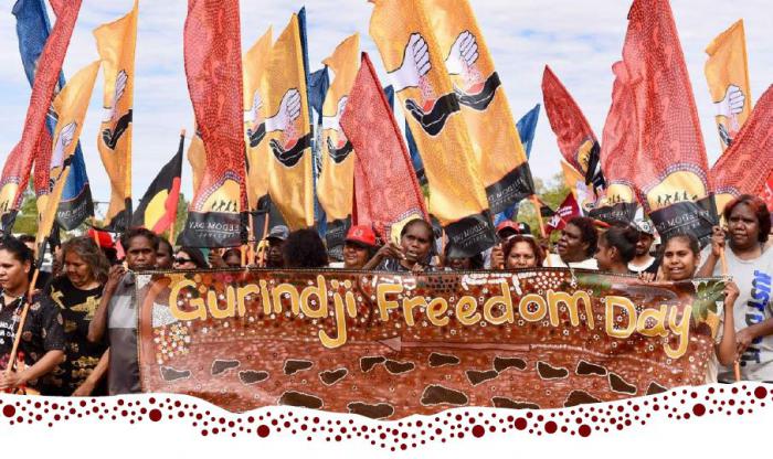 A group of people stand shoulder to shoulder holding up a banner which says Gurindji Freedom Day. Many behind them hold up flags featuring a white hand passing soil to a black hand.