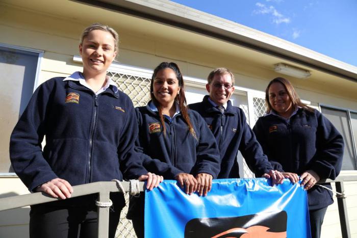 Four young adults wearing blue jumpers standing in front of a building and hanging onto a rail.