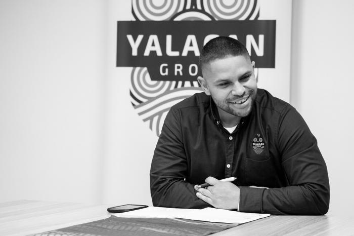 Young adult man in dark shirt sits at a table. In the background is a logo including the words Yalagan Group.