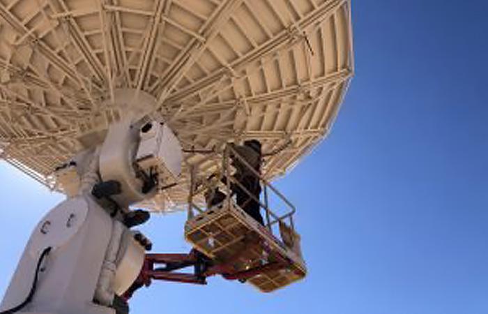 A man in workwear stands on a secure platform while he works below a large white satellite dish.