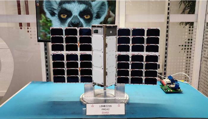 A mechanical device with white panelling and banks of black coloured photovoltaic cells on either side sits on a blue table. Behind, on the table, is an electronic device and in the background is a wall including a picture of a Lemur.