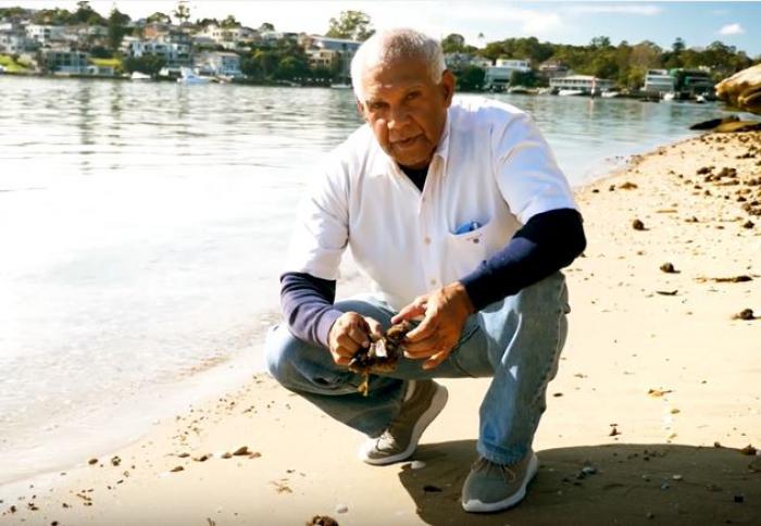 Elderly Aboriginal man in white shirt and jeans squats next to a river on a sandy shore. In the background on the other side of the river are houses and trees.