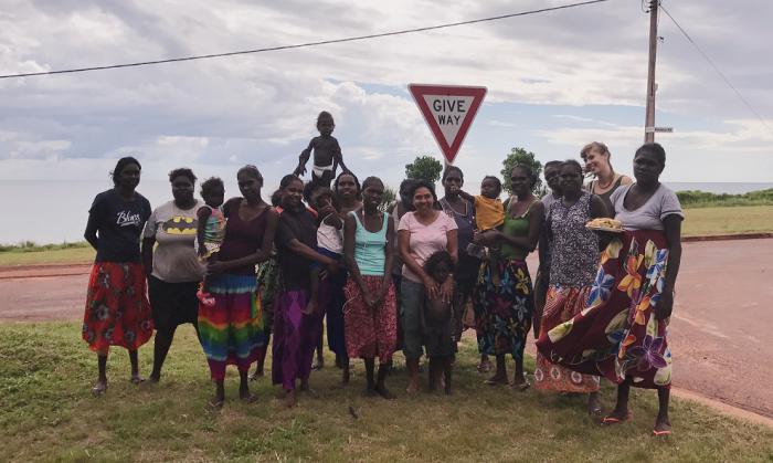 A group of mainly Aboriginal women and children dressed in an array of colourful clothing stand on grass next to a road corner with a telegraph pole in the background and a sign that says Give Way.